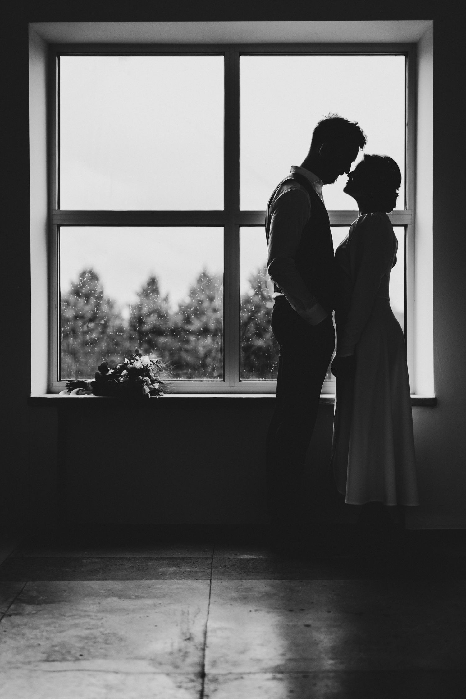 Black and white silhouette of a couple embracing romantically in front of a large window with trees outside.