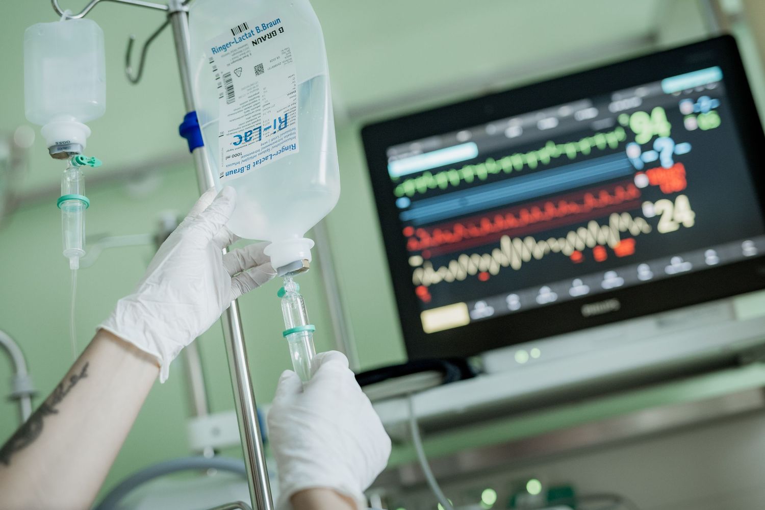 Gloved hands adjusting an IV drip bag on a pole beside a patient monitor displaying vital signs in a hospital ICU.