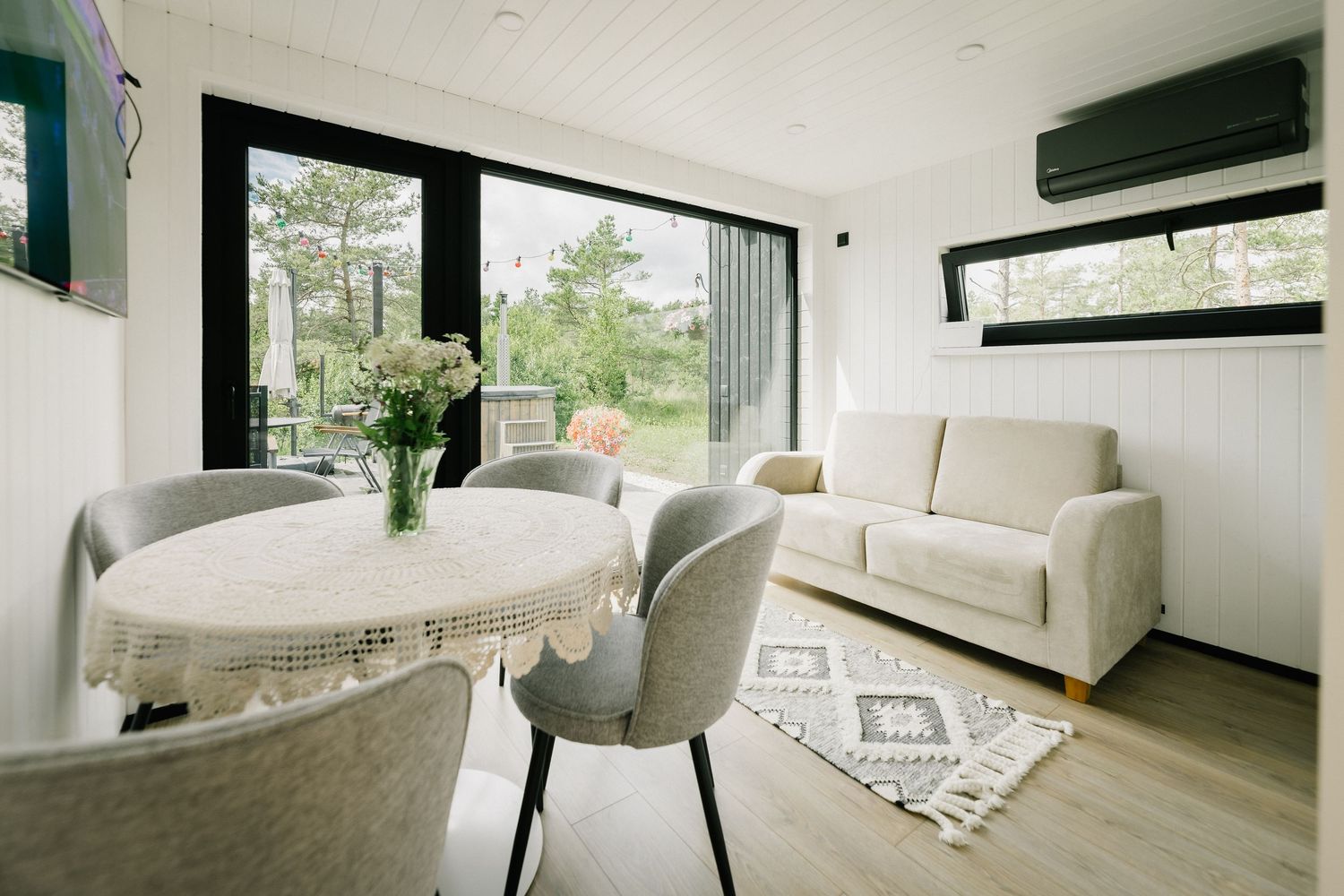 Bright tiny home interior with dining table, gray chairs, white sofa, patterned rug, and large windows overlooking greenery.