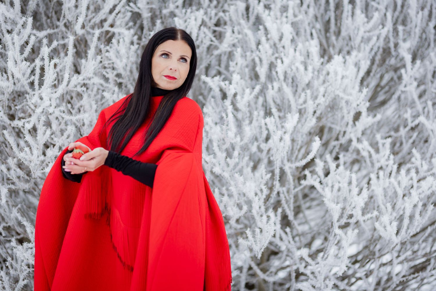 Woman in red cape coat poses before frost-covered white branches in a winter landscape.