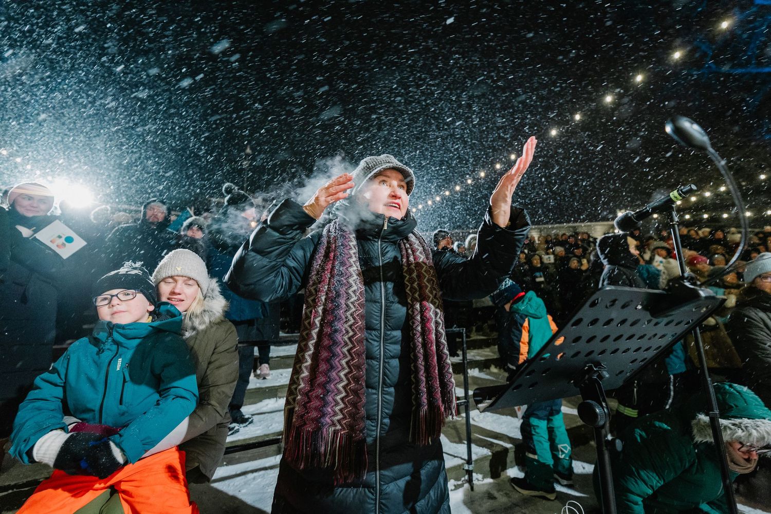 Conductor leads outdoor winter choir performance in snowfall, wearing scarf and winter jacket under bright stage lights.