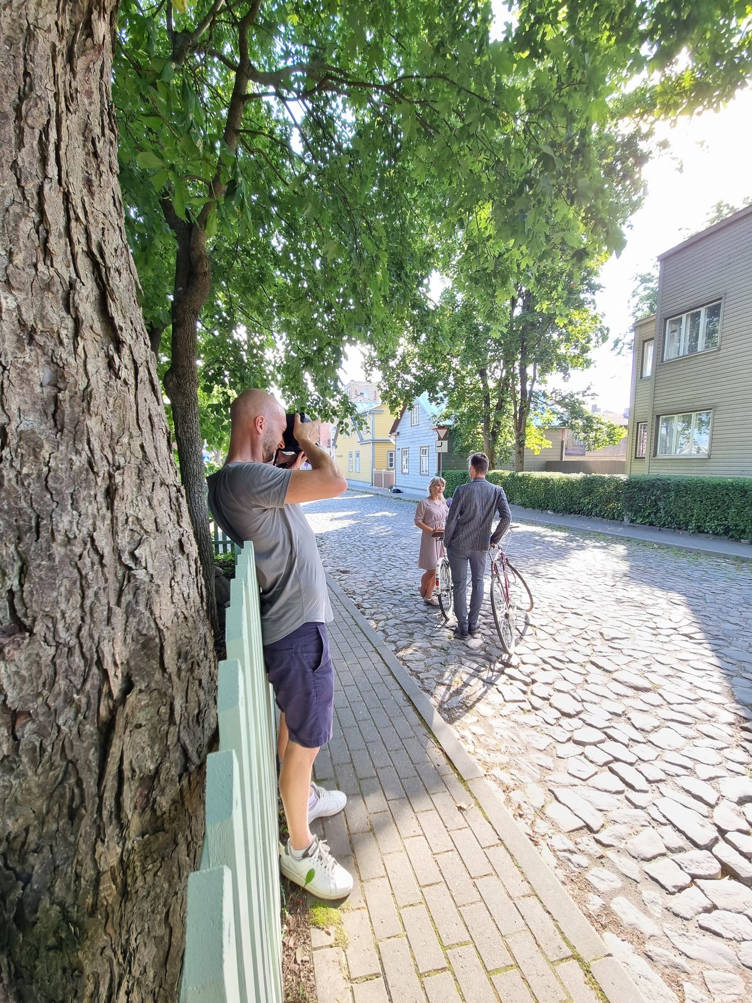 Photographer capturing a couple with a bicycle on a cobblestone street lined with green trees.