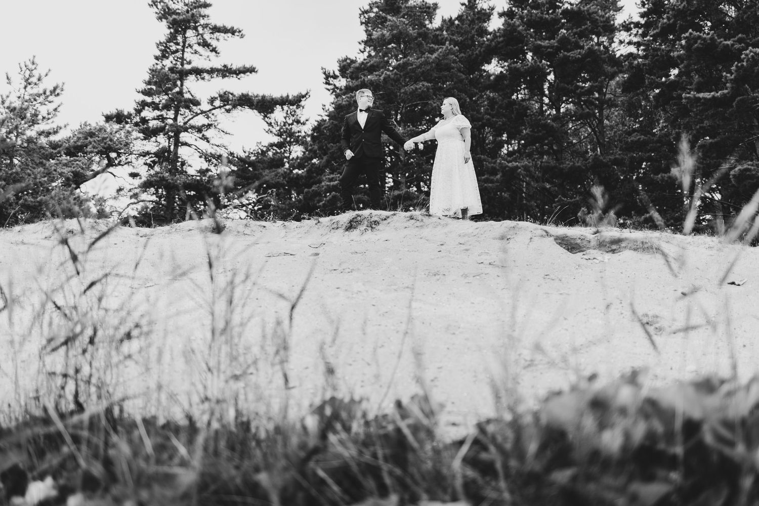Black and white photo of bride and groom holding hands on a sandy hill among tall grass.