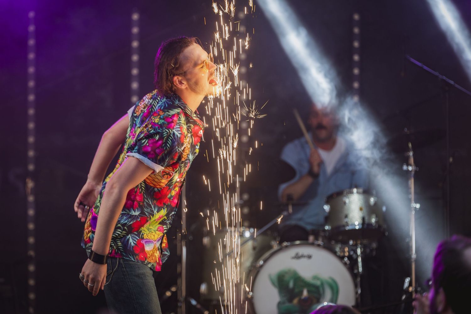 Rock band performing live on stage with pyrotechnic sparks, vocalist in floral shirt and drummer visible in background.