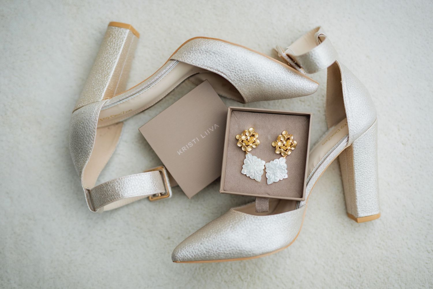 Elegant bridal flat lay with silver block heel shoes and gold floral earrings in a neutral gift box.