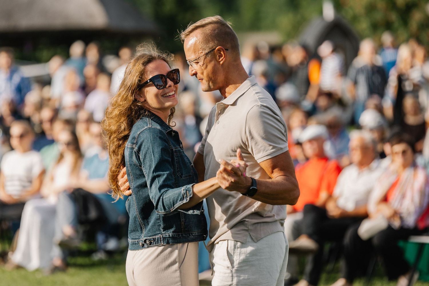 Couple dancing outdoors at a sunny event with a crowd of spectators seated in the background.