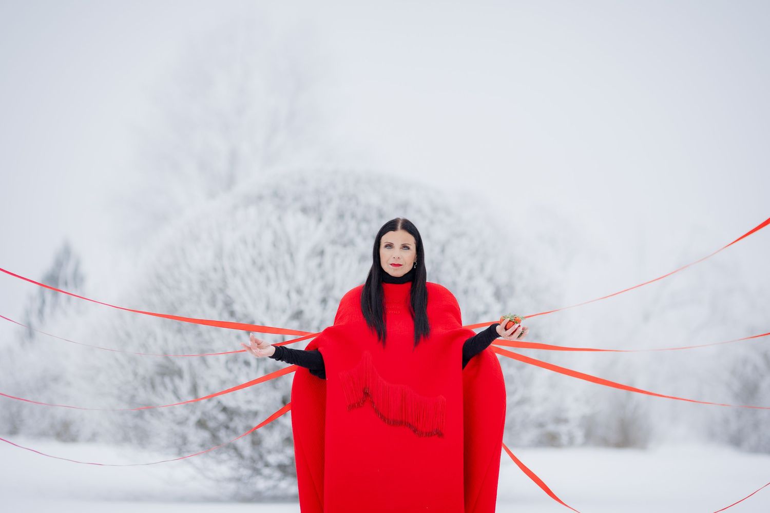 Woman in red cape holds red ribbons in snowy winter landscape with frost-covered trees behind her.