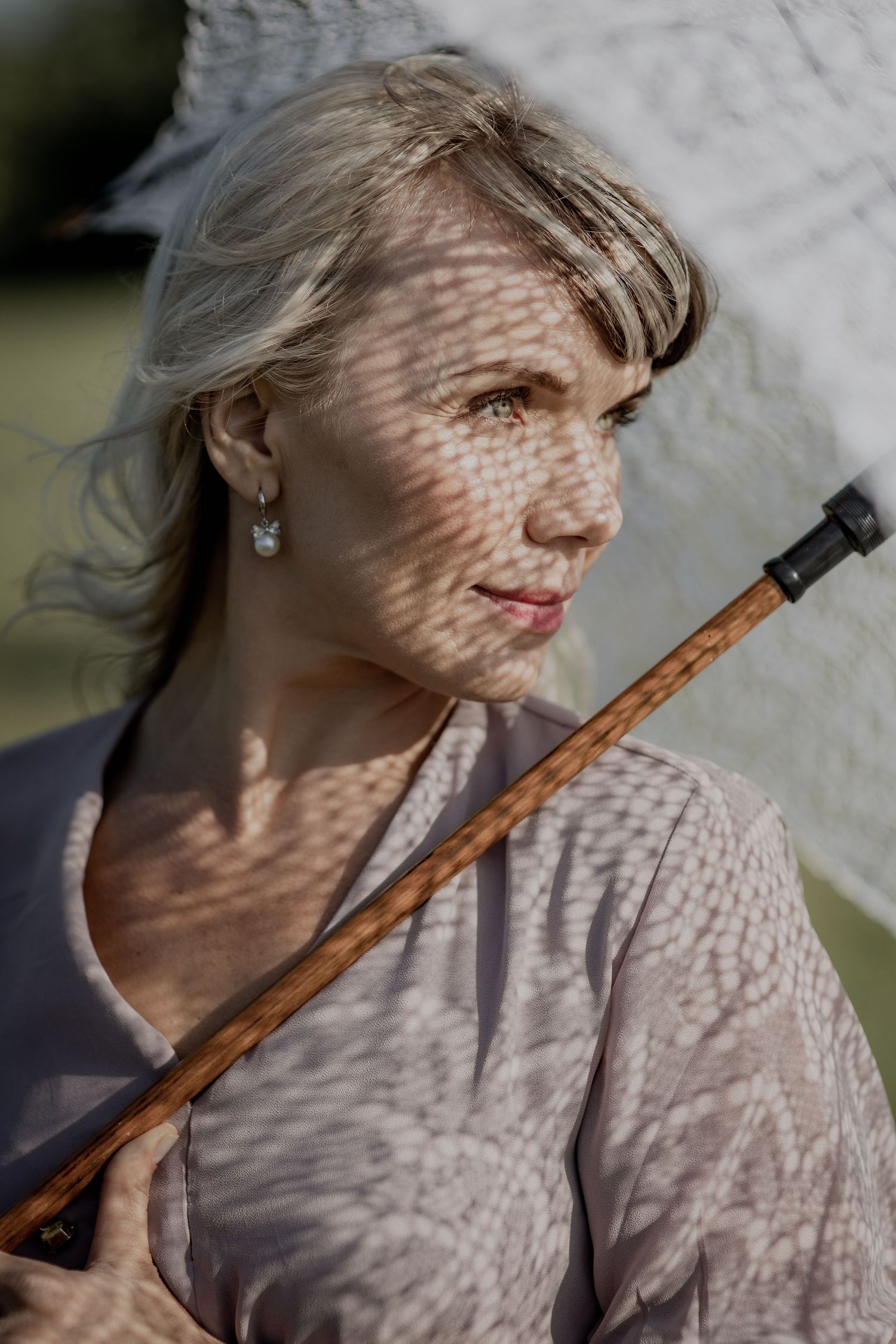 Mature woman with gray hair holding a parasol, dappled sunlight casting shadows on her face outdoors.