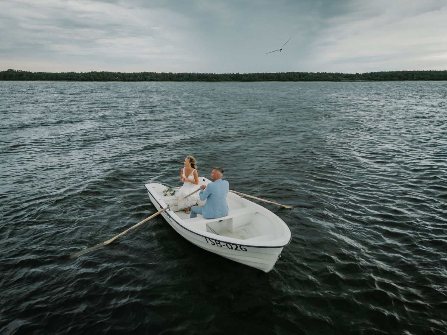 A couple in wedding attire sits romantically in a small white rowboat on a calm lake under a cloudy sky.