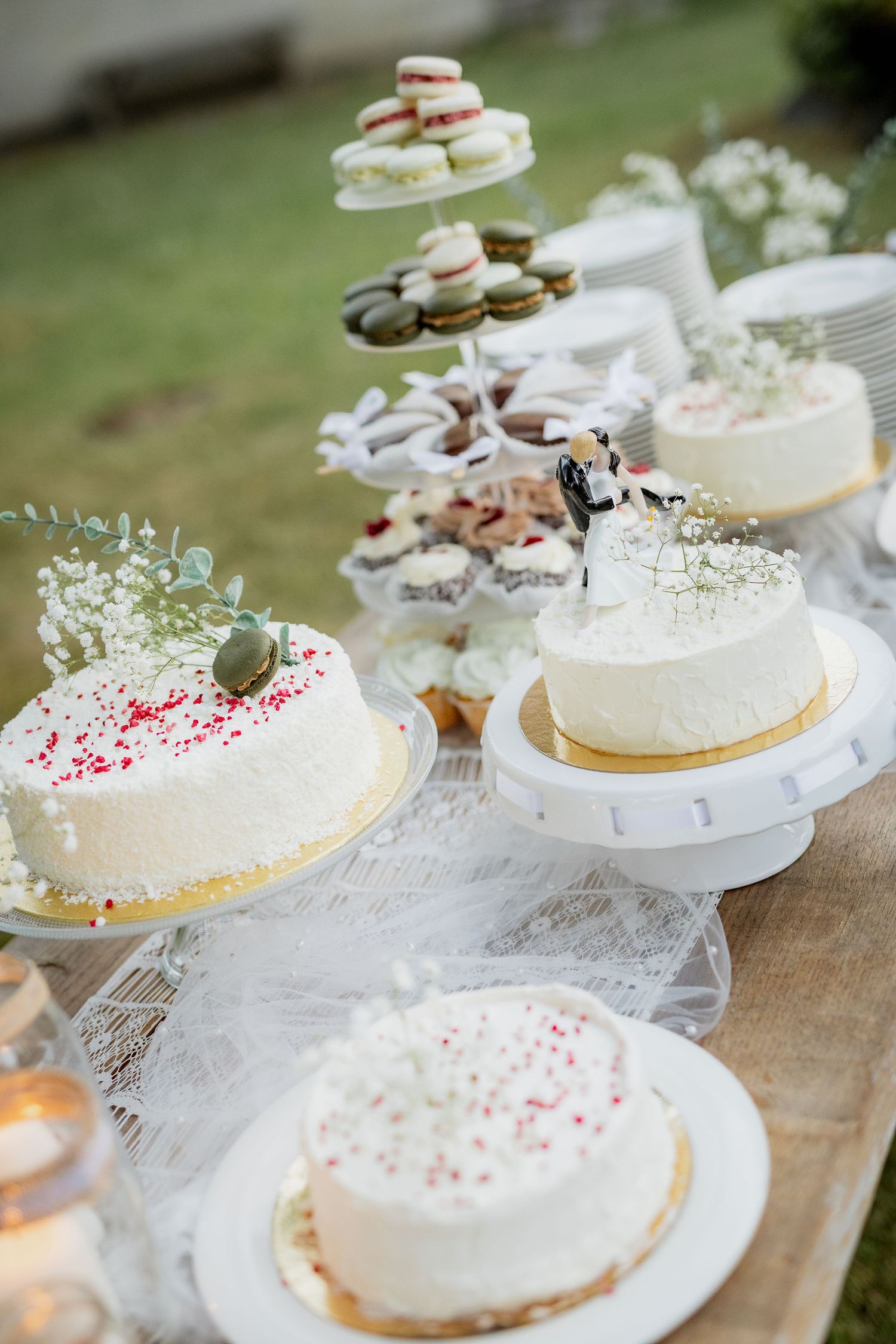 Elegant outdoor wedding dessert table with white frosted cakes, macarons on tiered stand, and bride and groom cake topper.