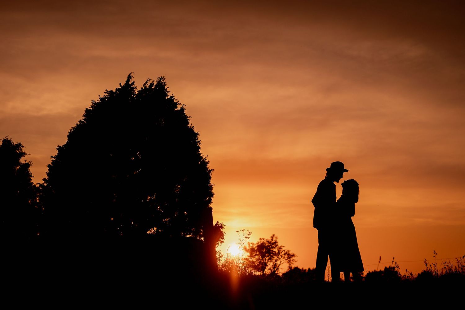 Silhouette of a romantic couple embracing at sunset with a warm orange sky and tree backdrop.