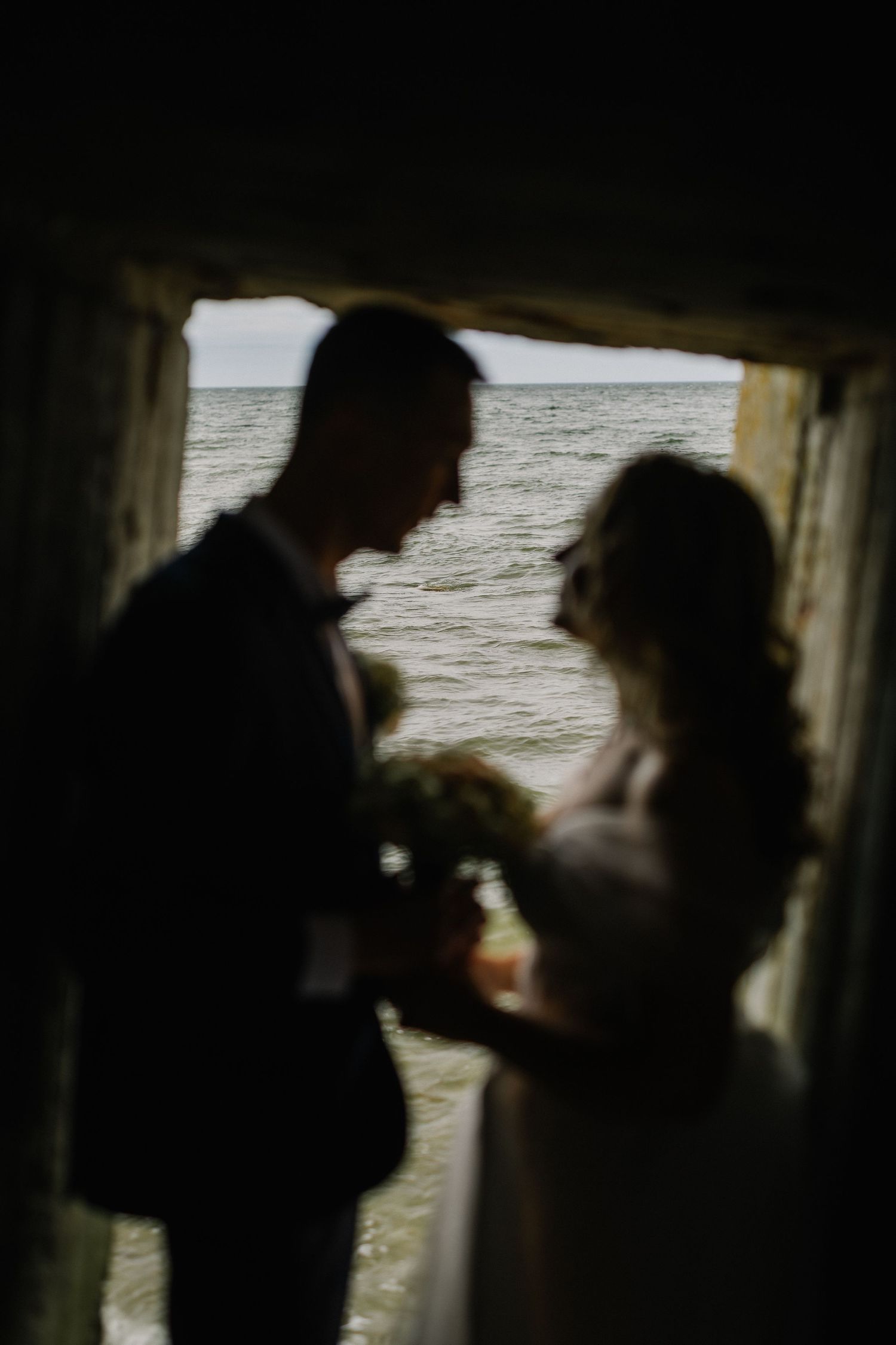 Silhouette of bride and groom facing each other with bouquet framed by dark archway and sea.