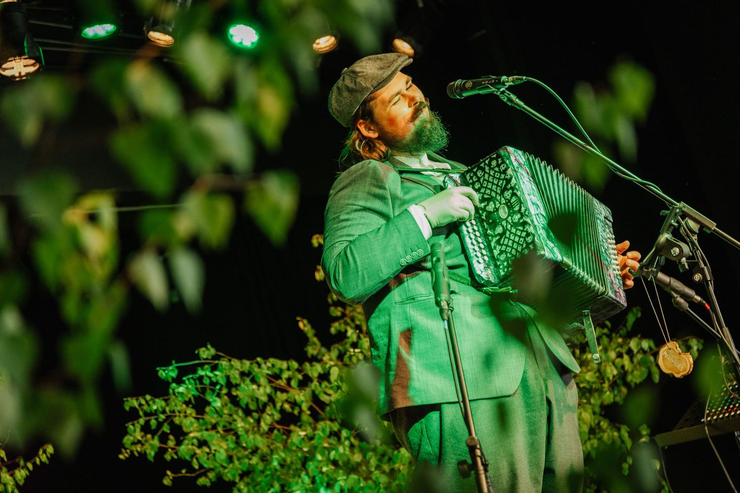 Bearded man in green suit playing accordion on stage surrounded by lush foliage under green lights.