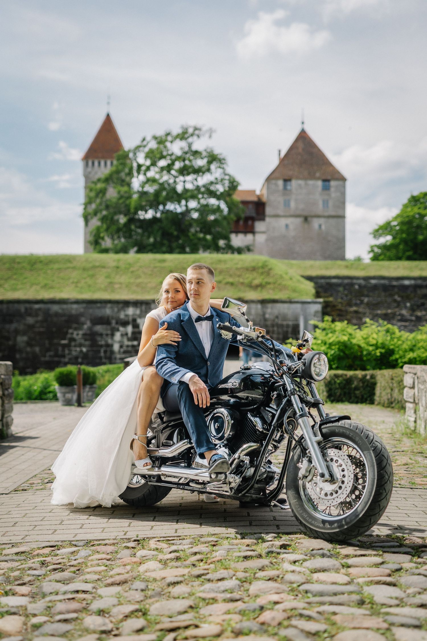 Bride and groom posing on a black motorcycle in front of a historic castle with towers on their wedding day.