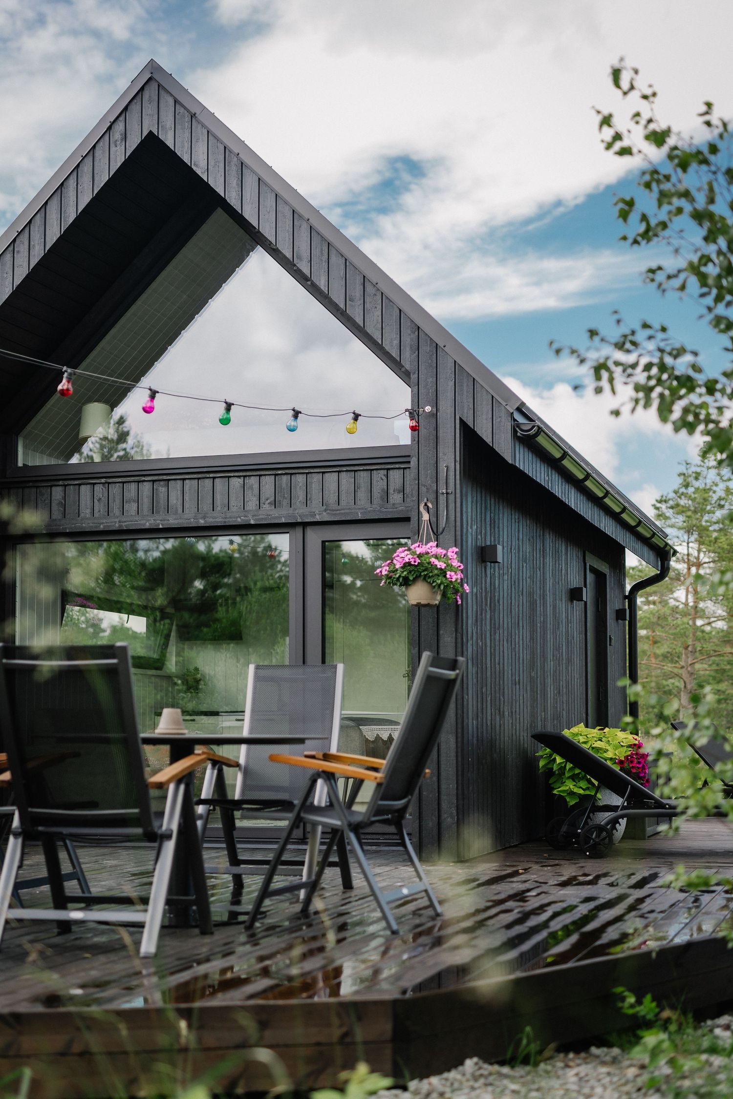 Dark wood cabin with gabled roof, string lights, outdoor patio furniture, and lush greenery on a cloudy day.