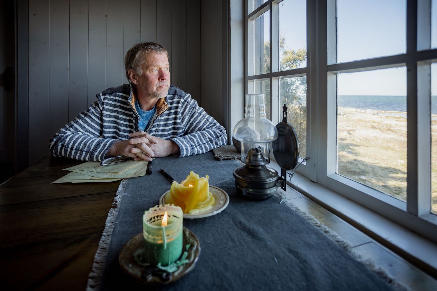 Middle-aged man in striped shirt sitting at table gazing through window at scenic ocean view.