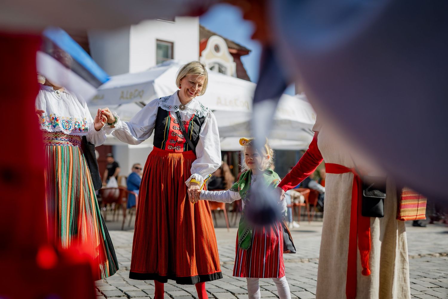 Woman in traditional folk costume dancing with a child in a sunny town square during a cultural festival.