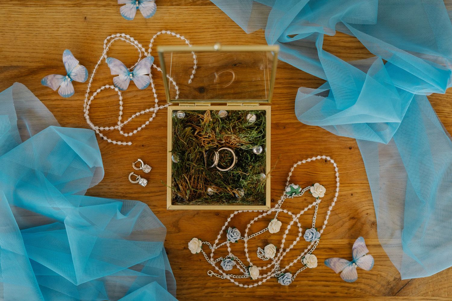 Wedding rings in a glass box surrounded by pearl necklaces, blue tulle, and butterfly decorations on a wooden surface.