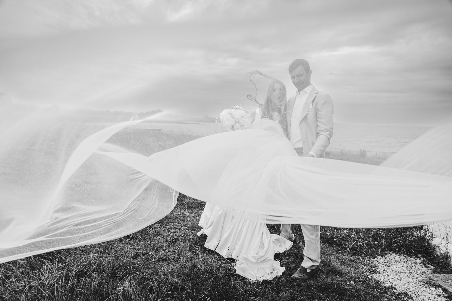 Black and white photo of bride and groom with wedding veil flowing dramatically in the wind by the ocean.