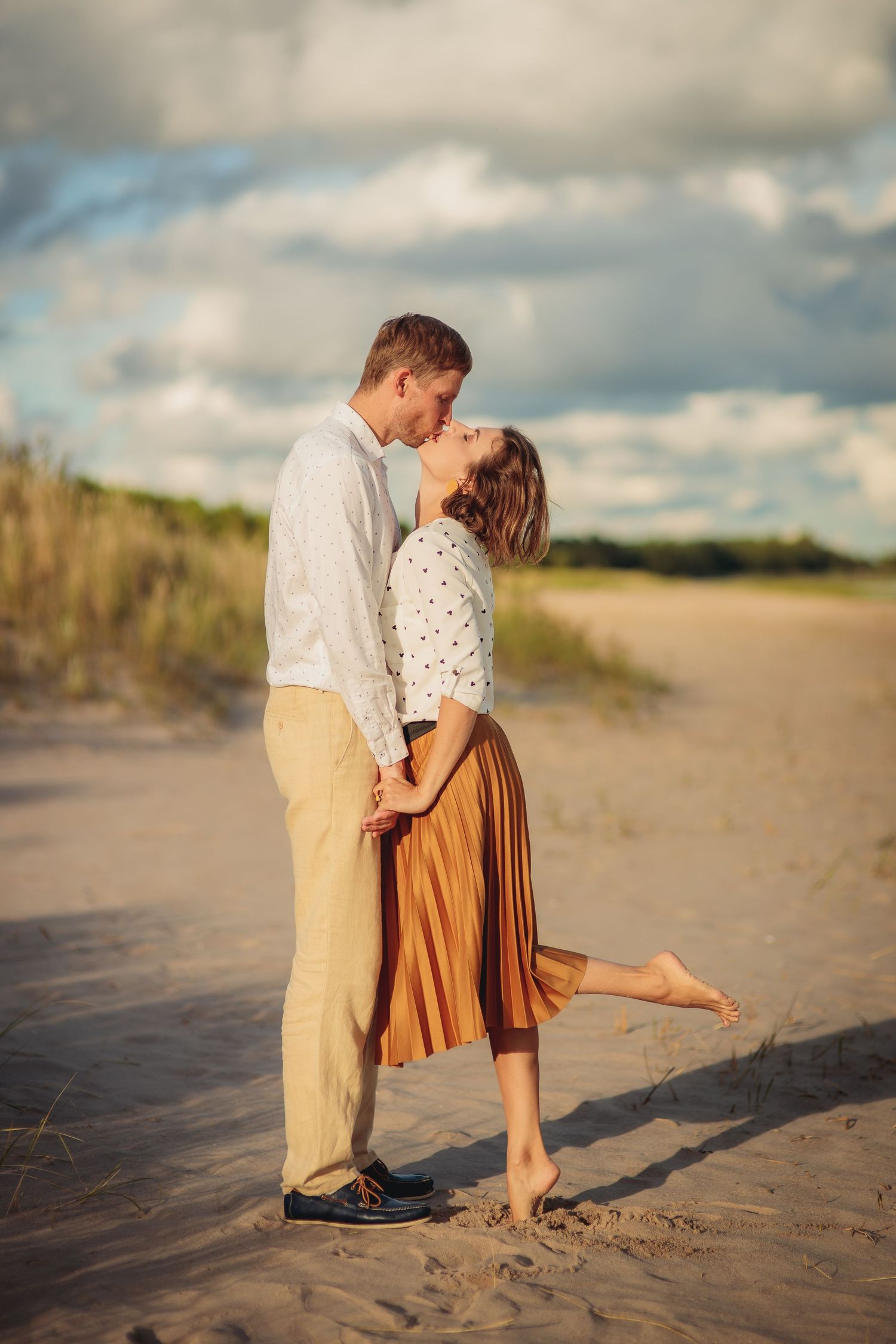 Romantic couple kissing on a sandy beach with cloudy sky, woman in brown skirt standing on tiptoes.