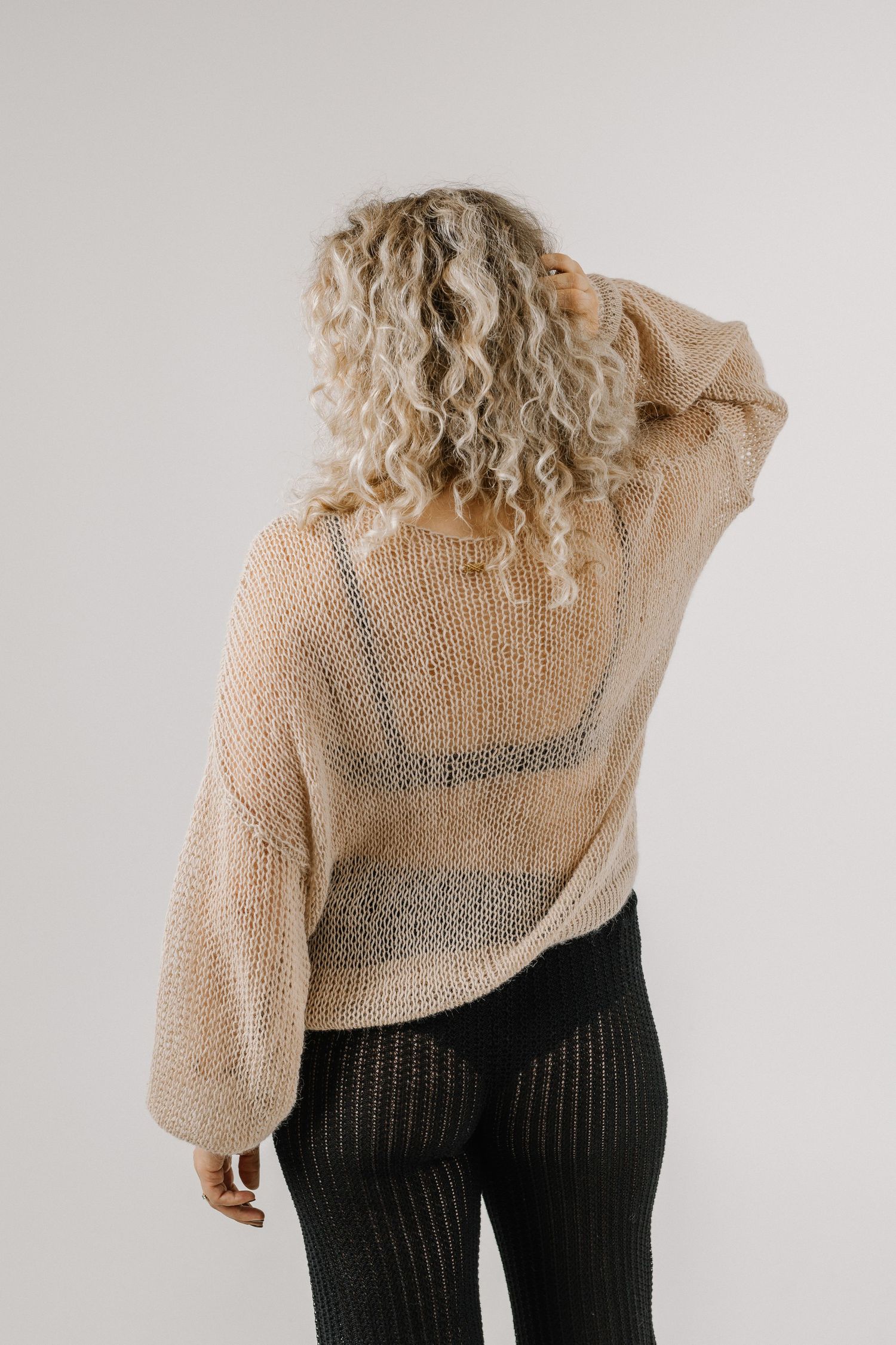 Woman in beige open-knit sweater and black ribbed pants, viewed from behind, with curly blonde hair.
