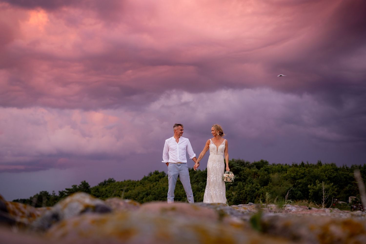 Bride and groom hold hands on rocky shore under dramatic pink and purple sunset sky.