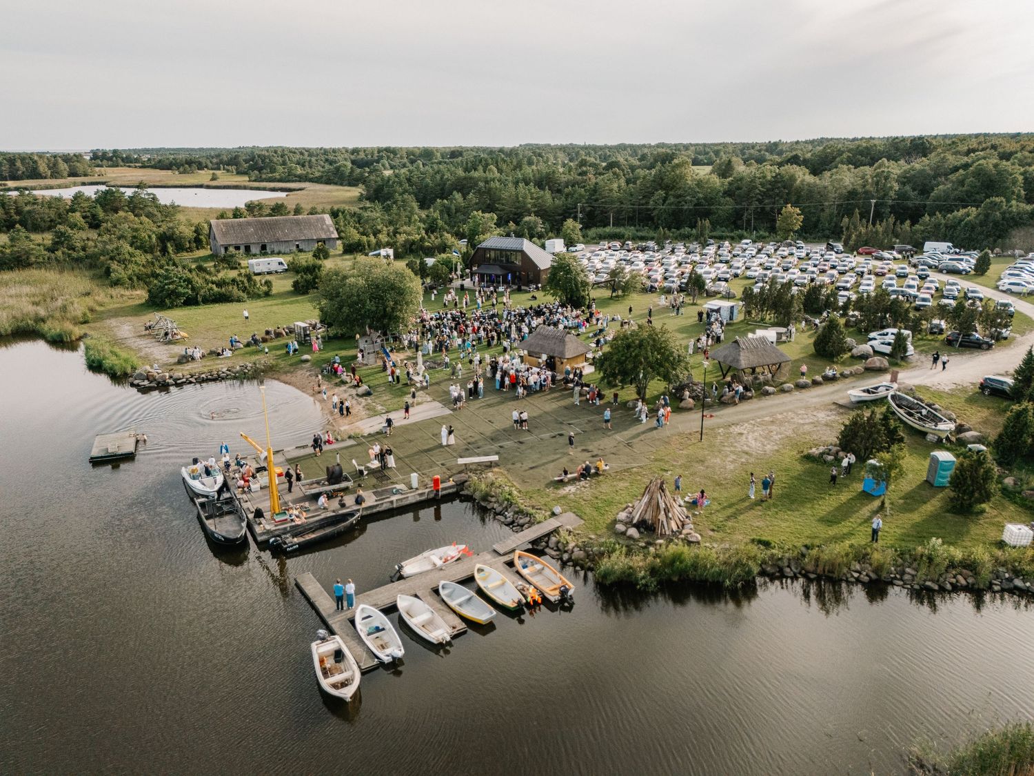 Aerial view of a waterfront festival with boats docked along a river, crowds gathering near a stage, and camping areas surrounded by trees.