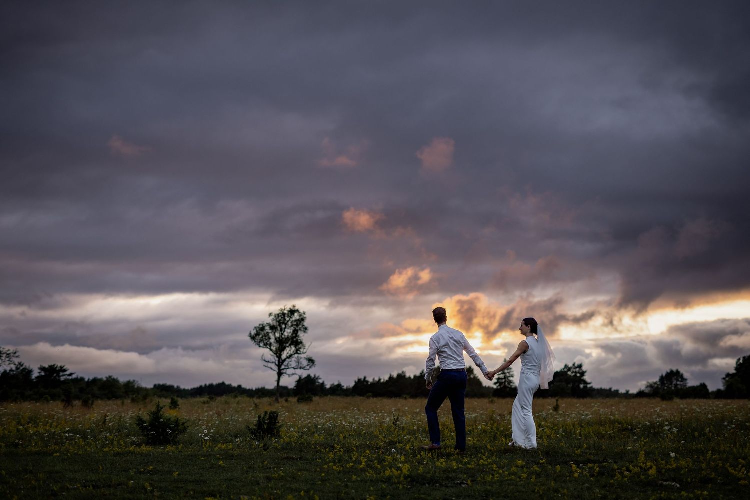 Bride and groom hold hands in a field at sunset under dramatic stormy clouds in a romantic outdoor wedding photo.