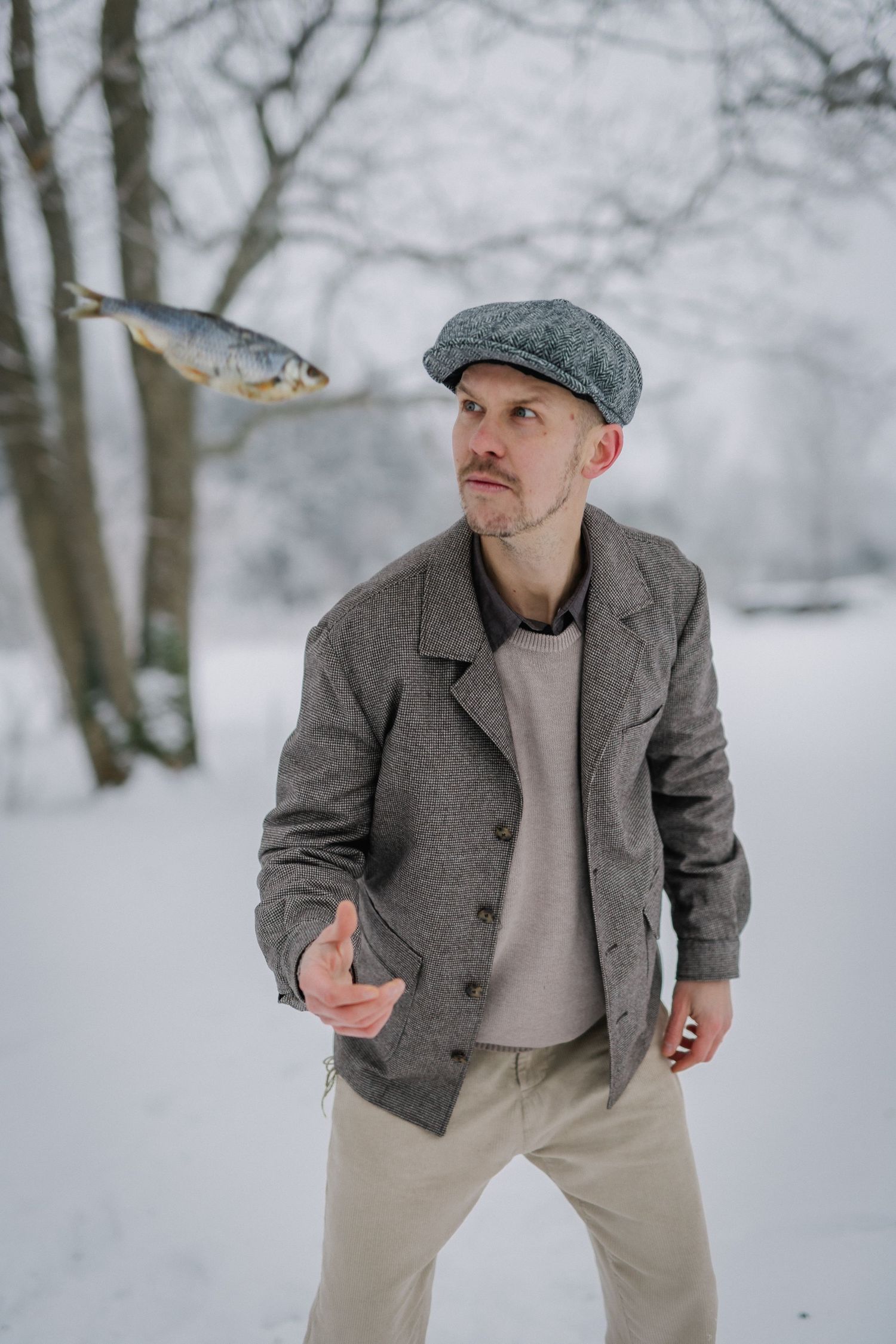 Man in gray flat cap and jacket tossing a fish in a snowy winter park setting.