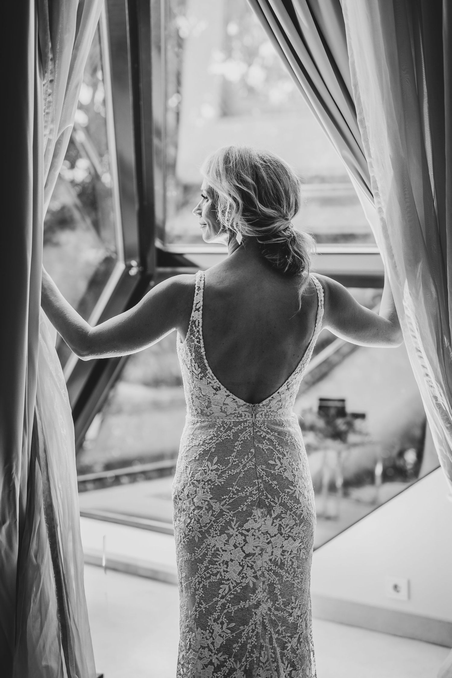Black and white photo of a bride in a backless lace wedding gown standing at an open window with curtains.