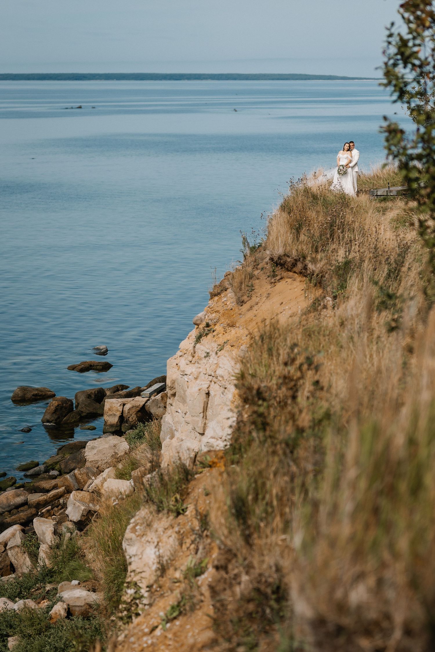 A couple in wedding attire stands on a rocky cliff overlooking a calm blue sea on their special day.