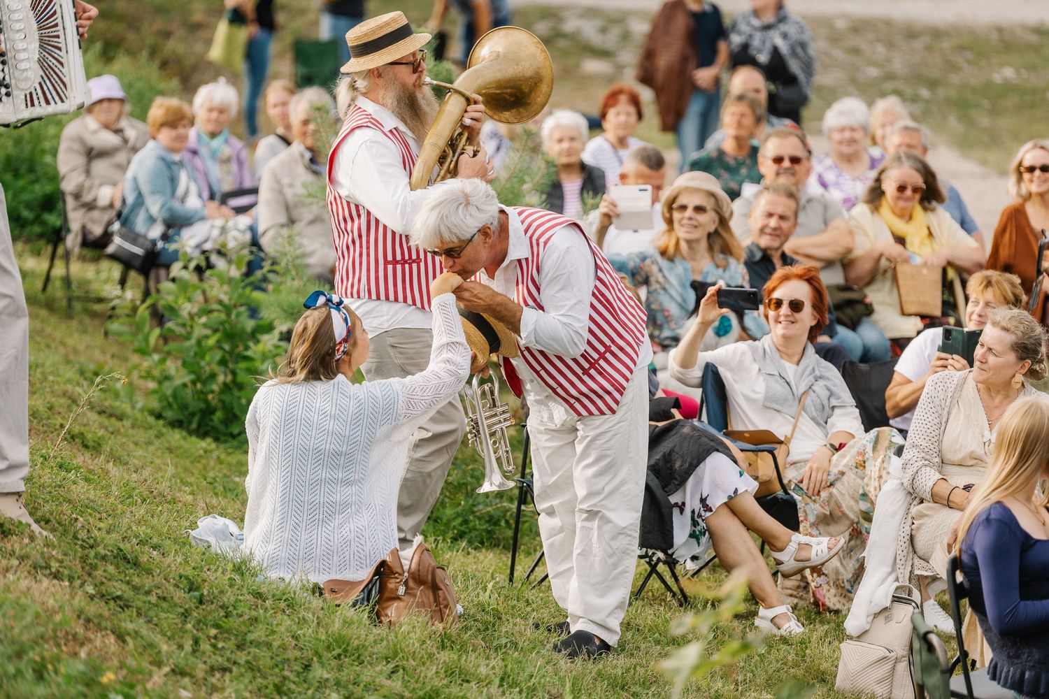 Folk musicians in red striped vests perform outdoors for a seated crowd at a traditional cultural festival.