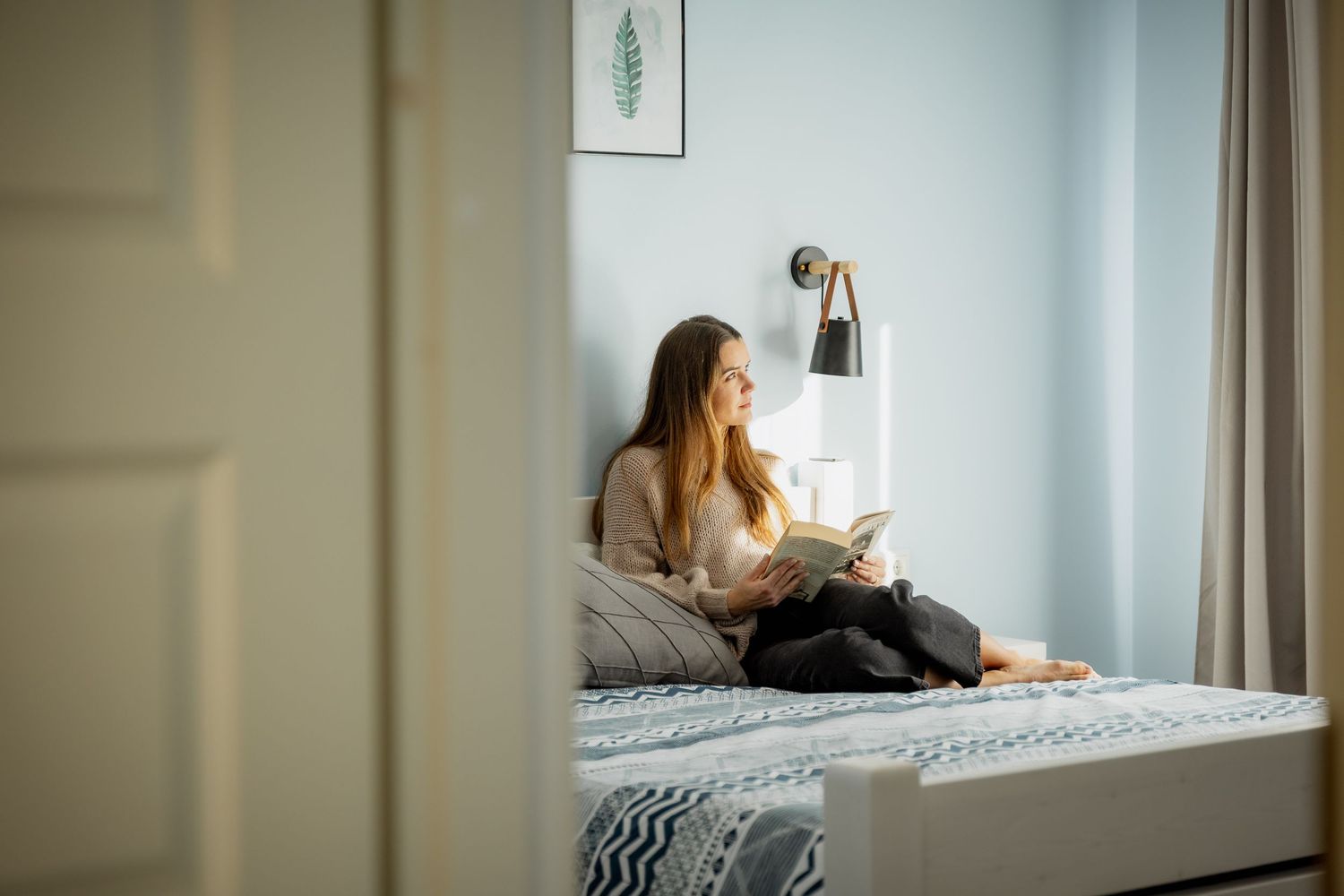 Young woman relaxing on bed reading a book in a cozy light blue bedroom with wall lamp and decorative art.