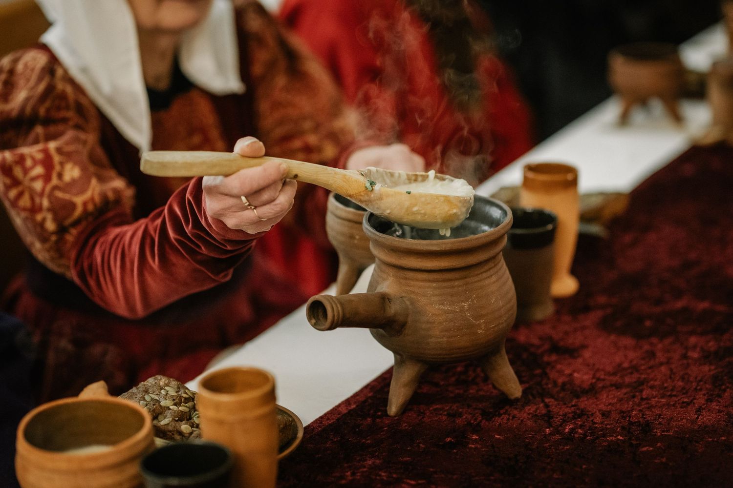 Person pouring ingredients into a clay pot during a traditional tea ceremony with wooden cups on a red cloth.