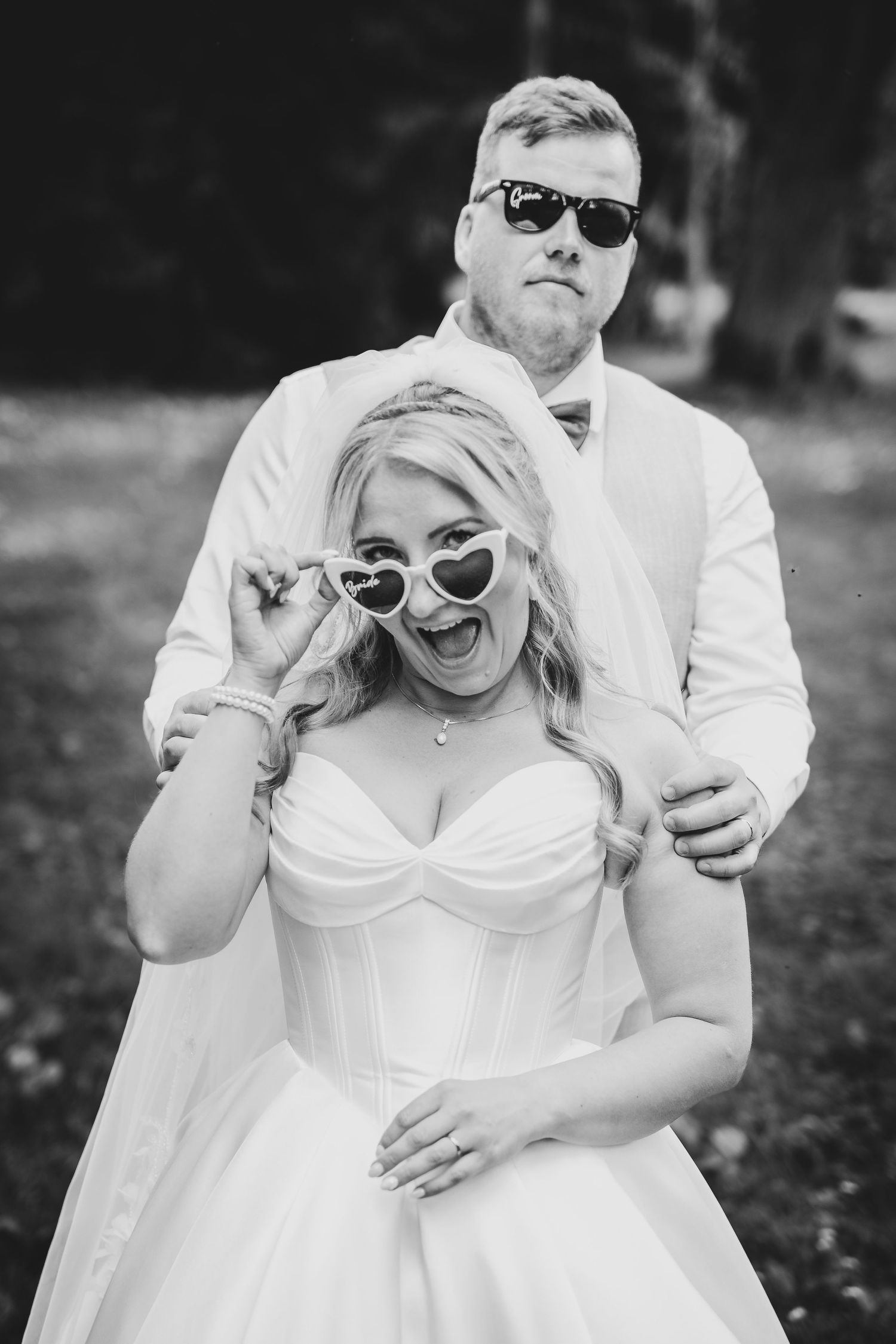 Black and white photo of a laughing bride in heart sunglasses and groom in shades posing outdoors on their wedding day.
