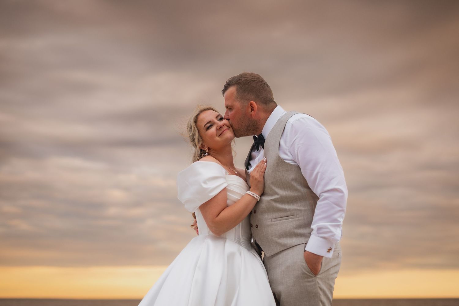 A bride and groom share a romantic moment at sunset on a beach, with dramatic clouds in the background.