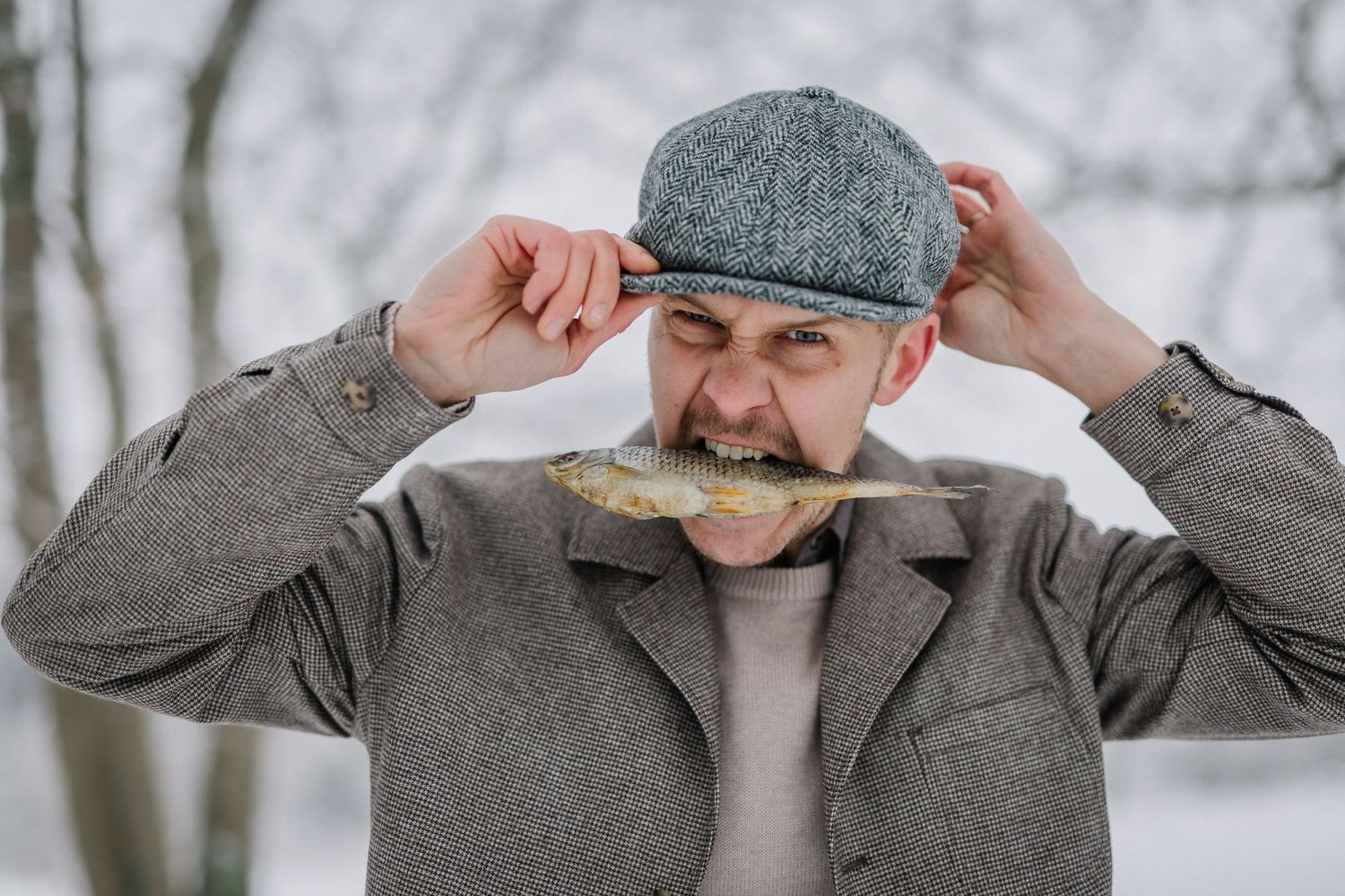 Man in gray coat and flat cap biting into a piece of bread outdoors in a snowy winter setting.