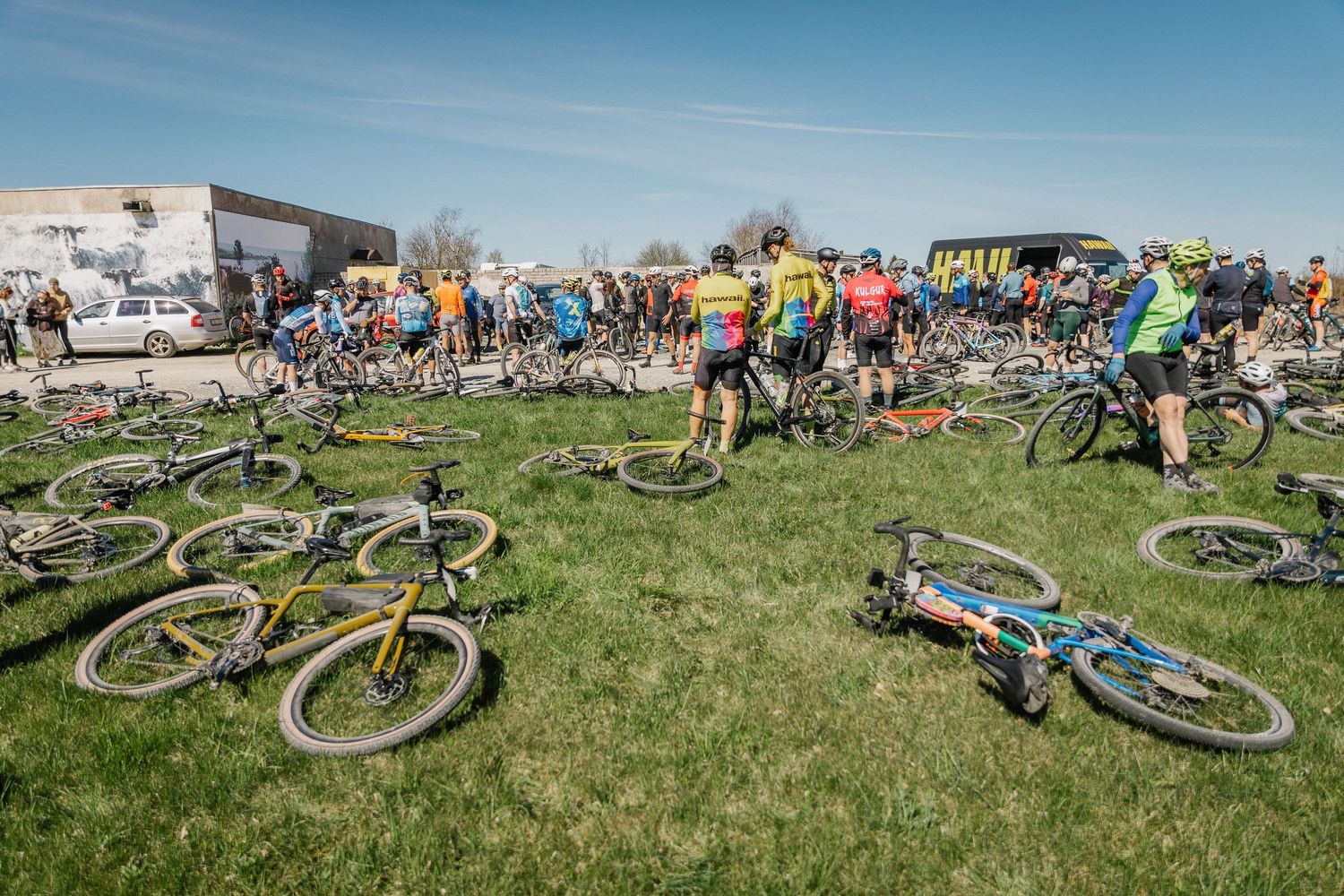 A large group of cyclists gather on a grassy field with mountain bikes laid on the ground at an outdoor cycling event.