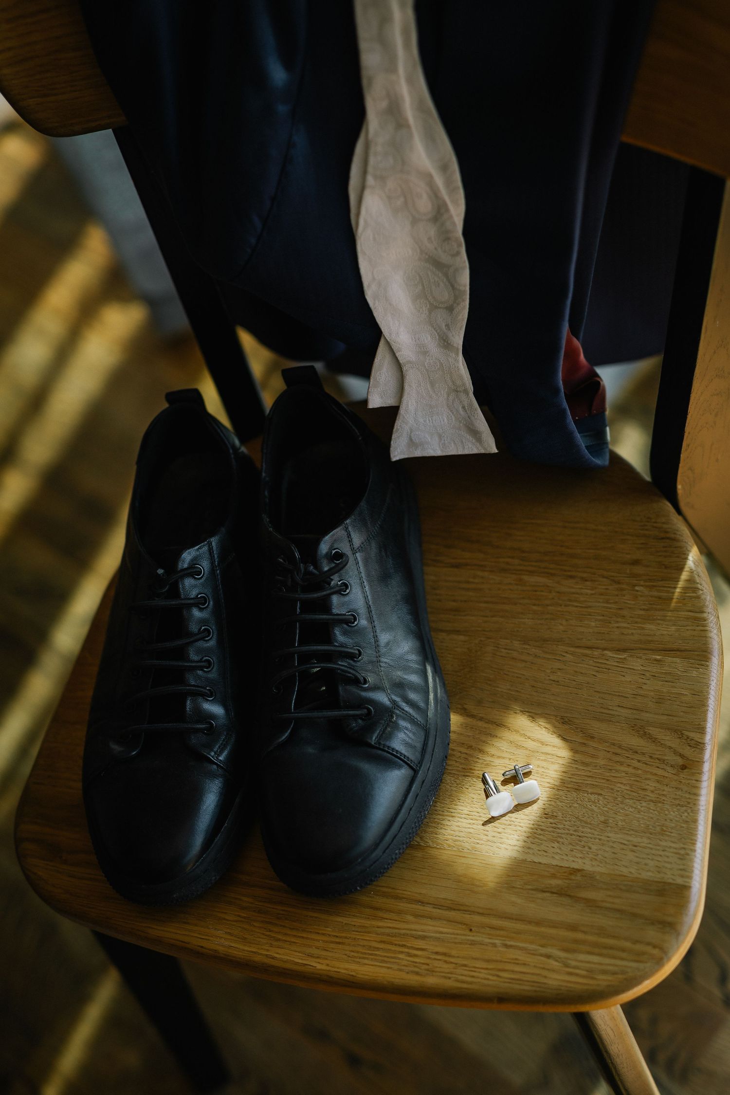 Black leather sneakers with laces on a wooden chair beside cufflinks and hanging suit jacket.