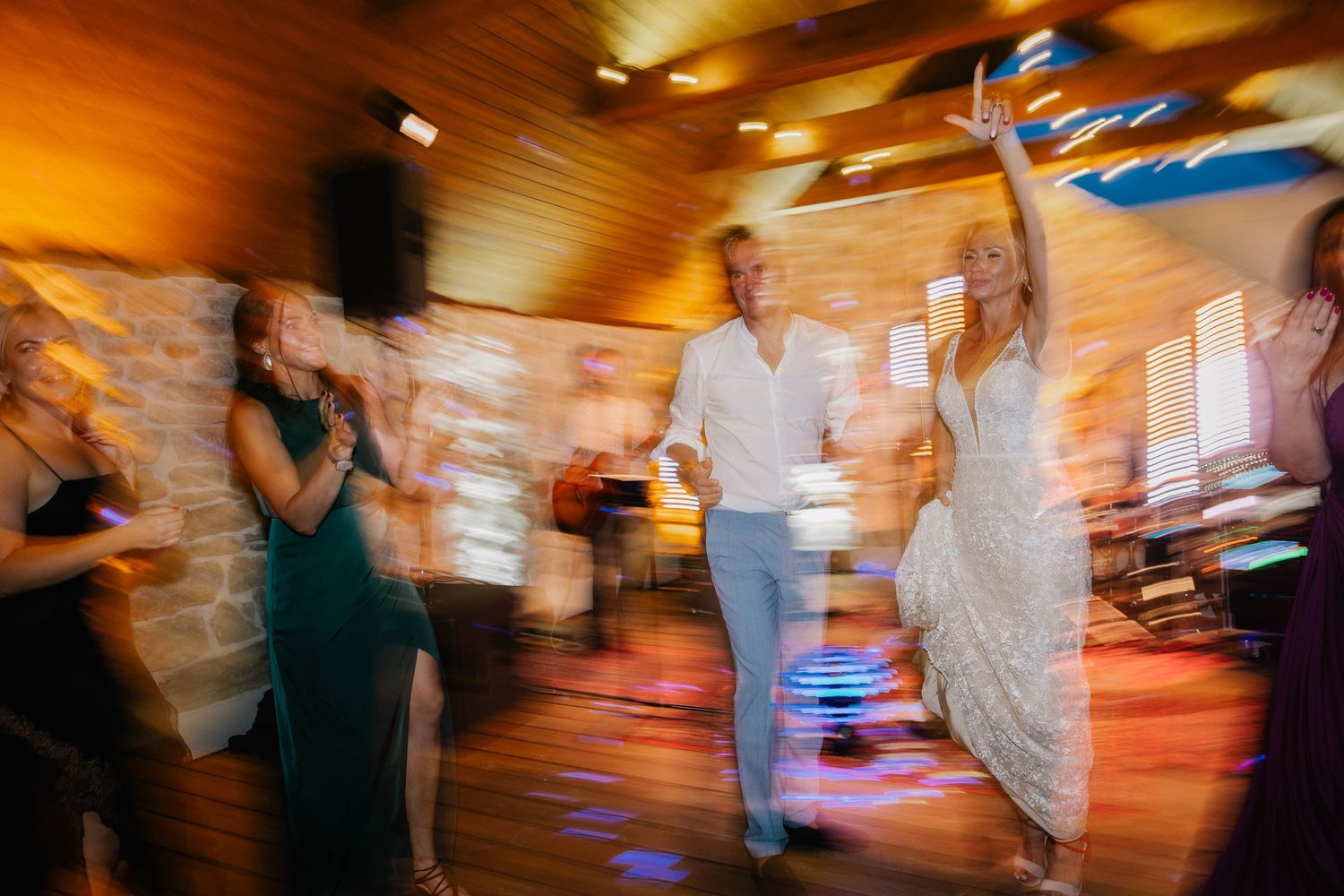 Bride and groom dancing joyfully with guests at a rustic wedding reception with warm lighting.