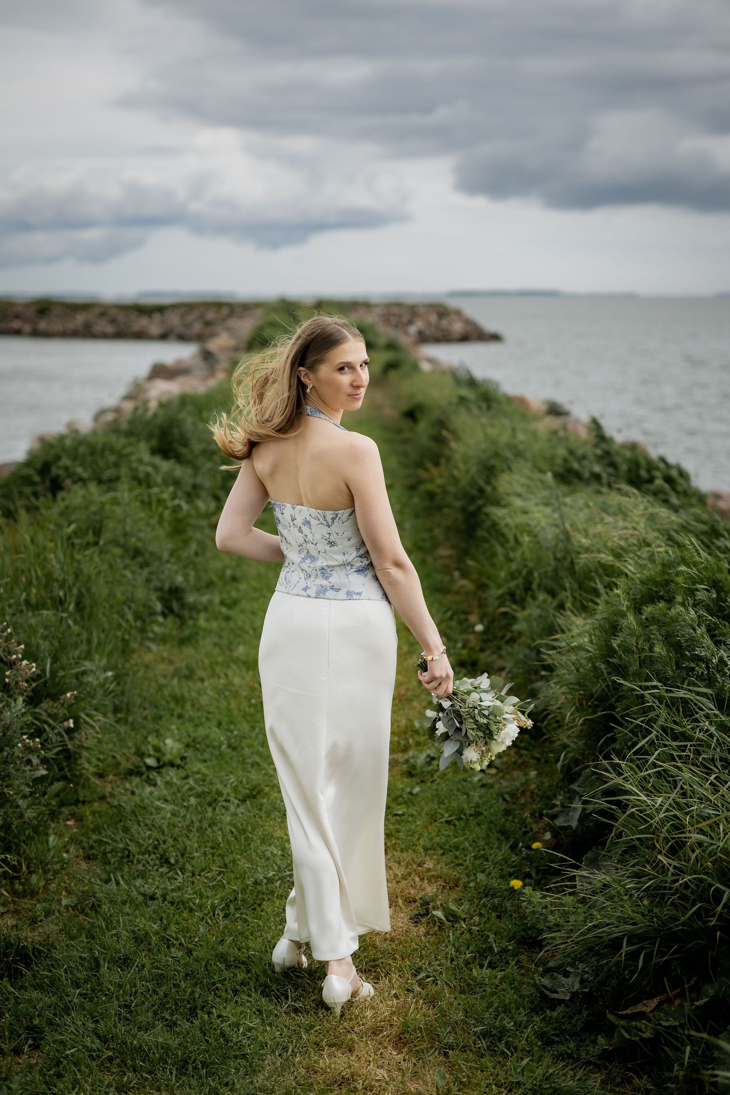 Bride in white skirt and floral corset holds bouquet on coastal path with rocky shoreline and cloudy sky.