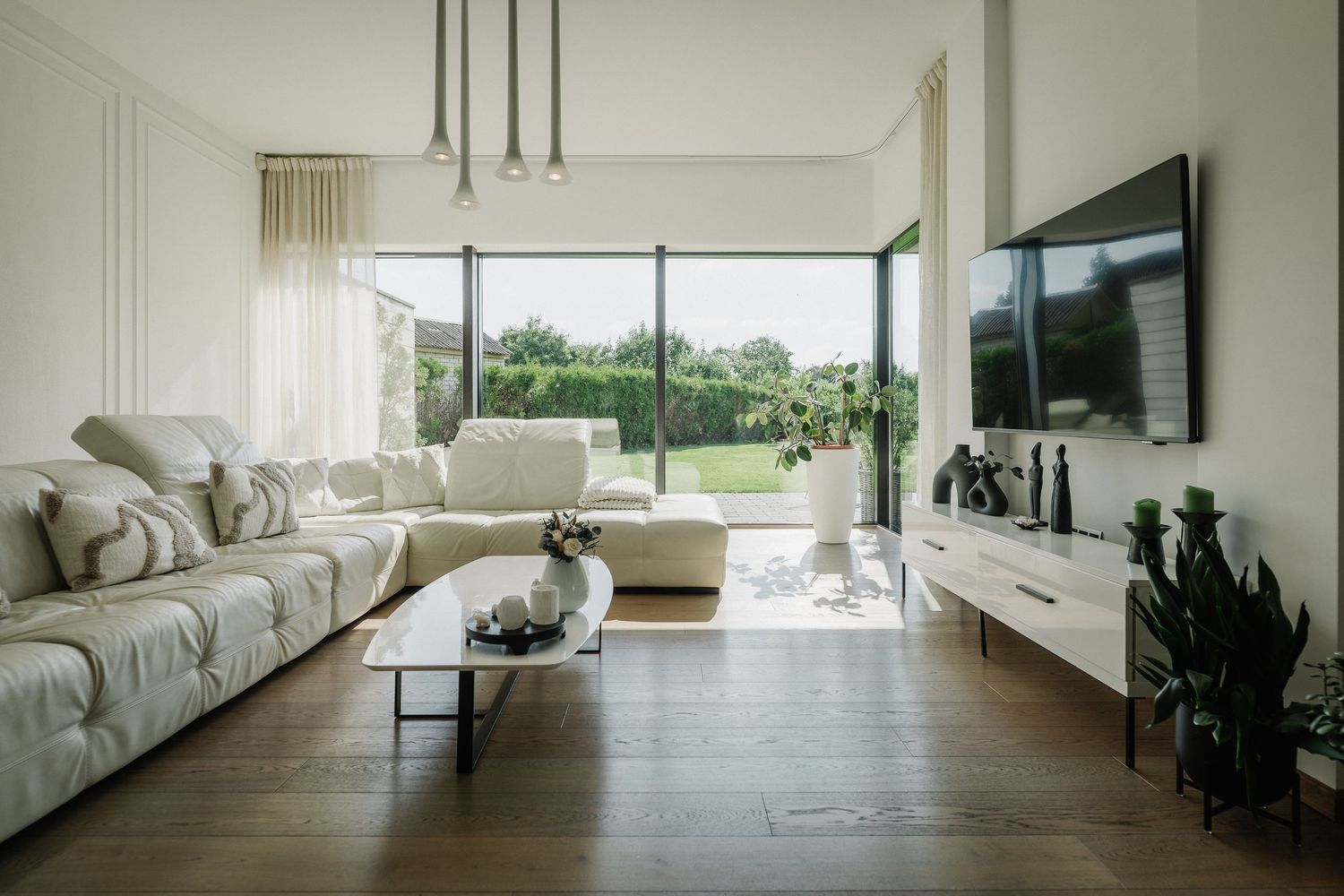 Modern minimalist living room with white sofas, hardwood floors, large glass doors, wall-mounted TV, and lush garden view.