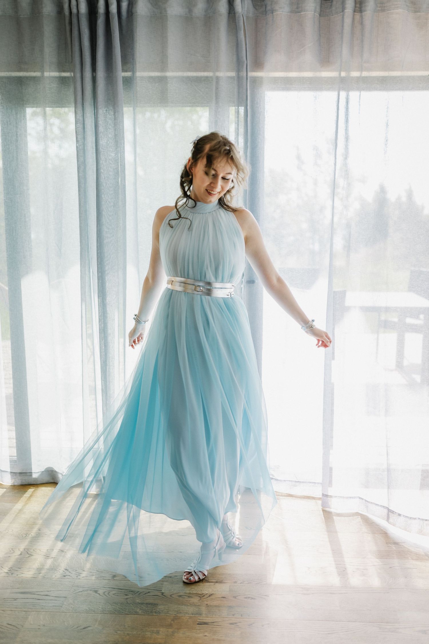 A woman in a flowing light blue tulle gown with silver belt poses gracefully by sheer curtains in a bright room.