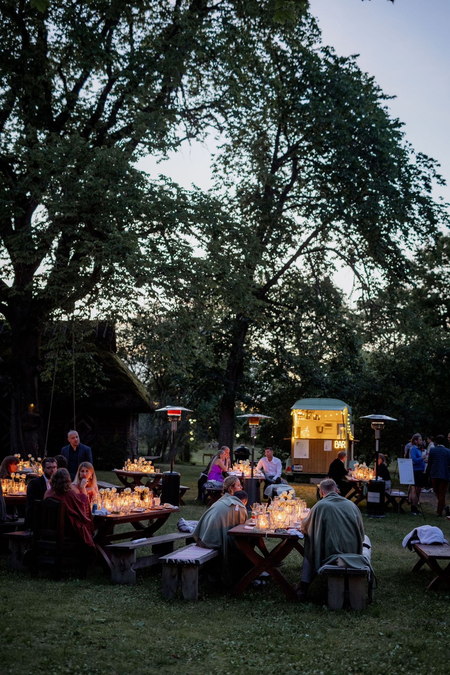 Outdoor evening gathering with guests dining at candlelit picnic tables under large trees near a glowing food truck.