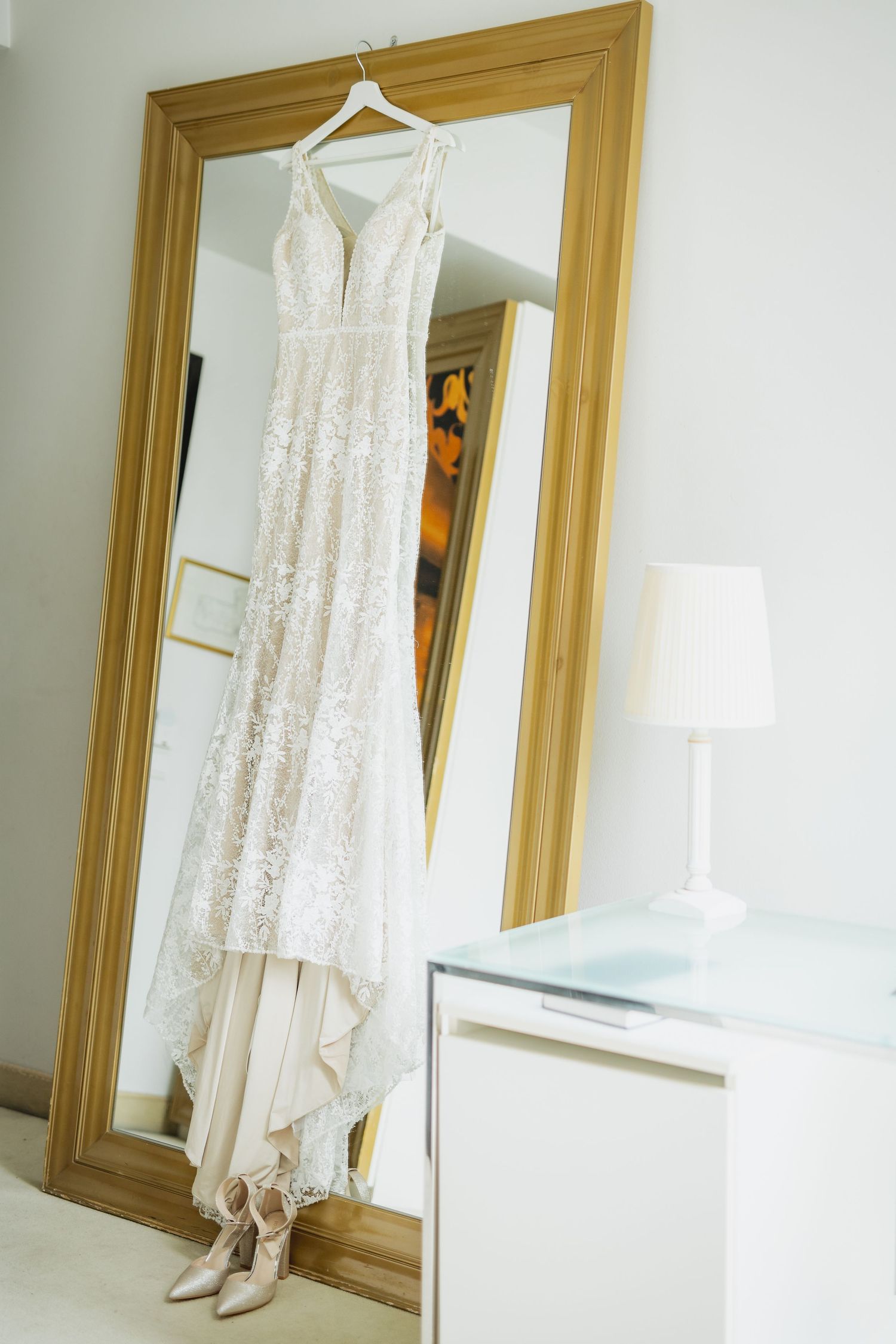 Elegant lace wedding dress hanging on a gold-framed mirror beside nude heels and a white bedside table with lamp.