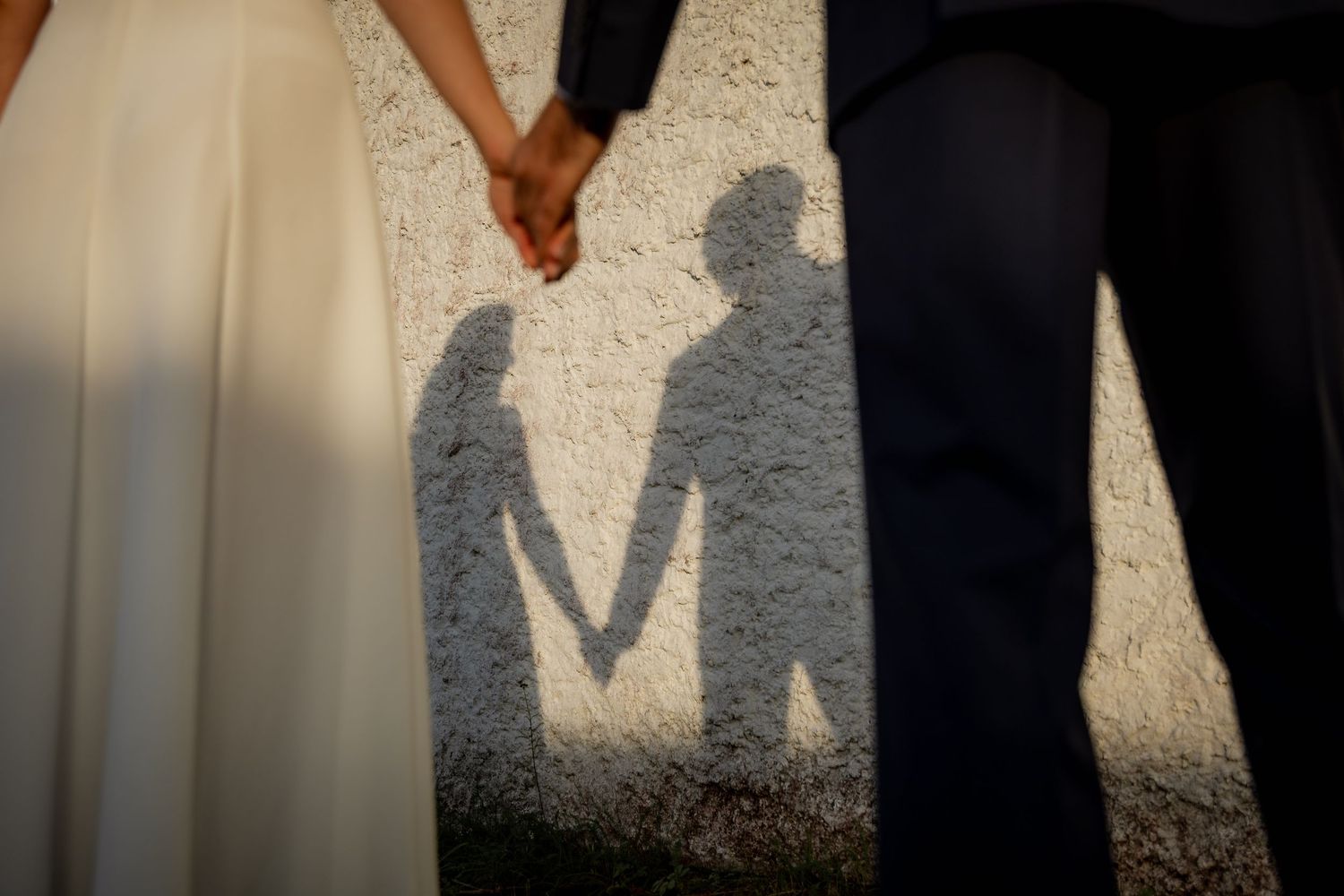 Couple holding hands casting romantic shadows on a sunlit wall in wedding attire.