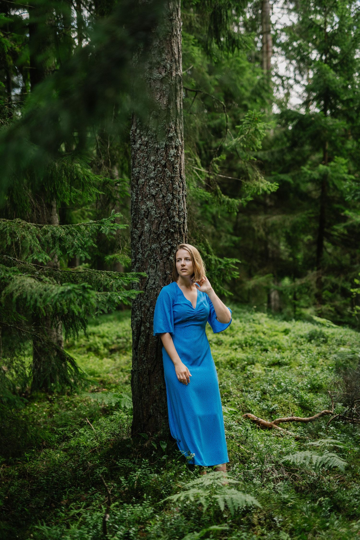 Blonde woman in elegant blue maxi dress leans against tree trunk in lush green forest.