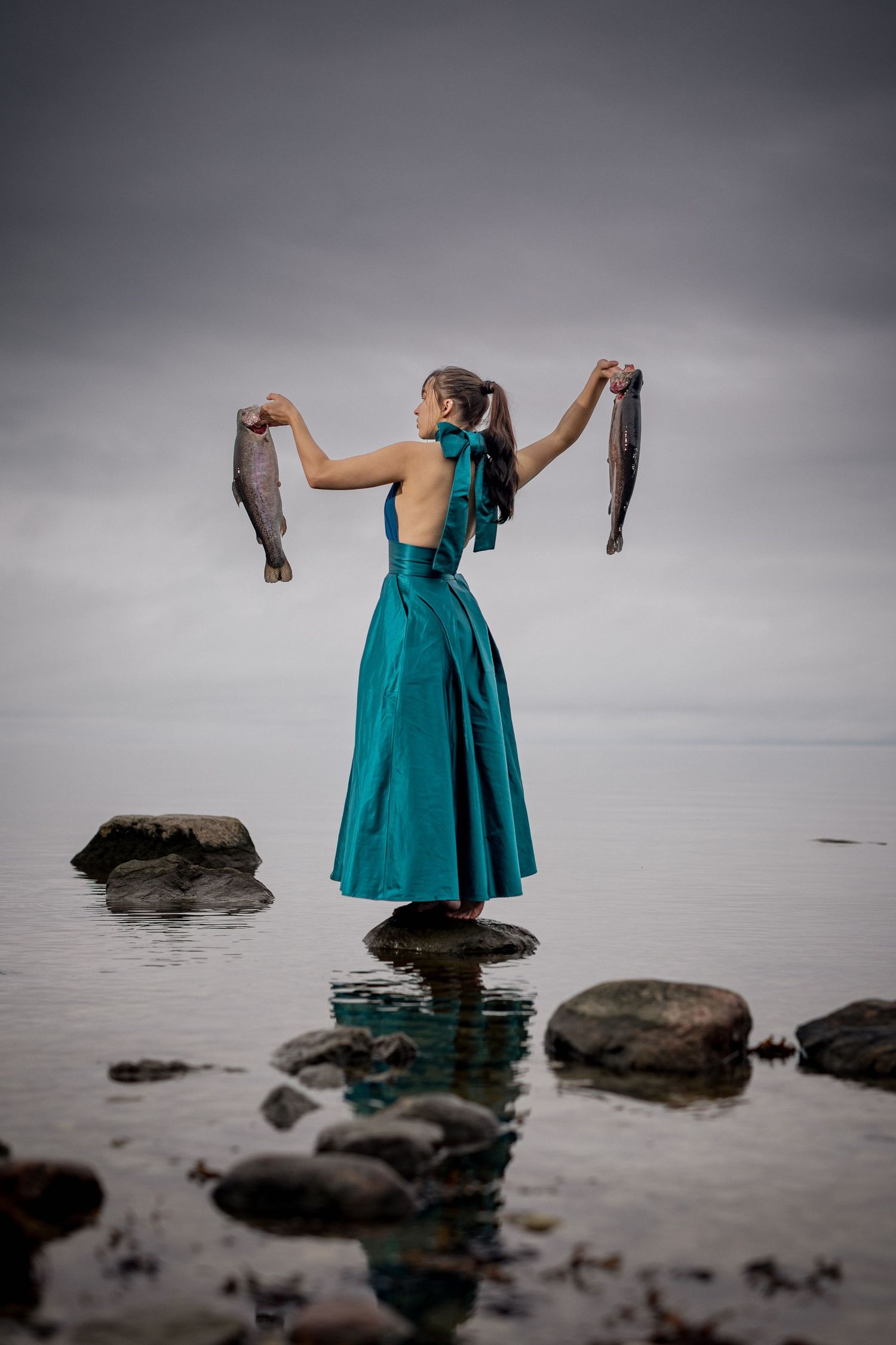 Woman in teal gown holding two fish aloft while standing on rocks in shallow water under overcast skies.