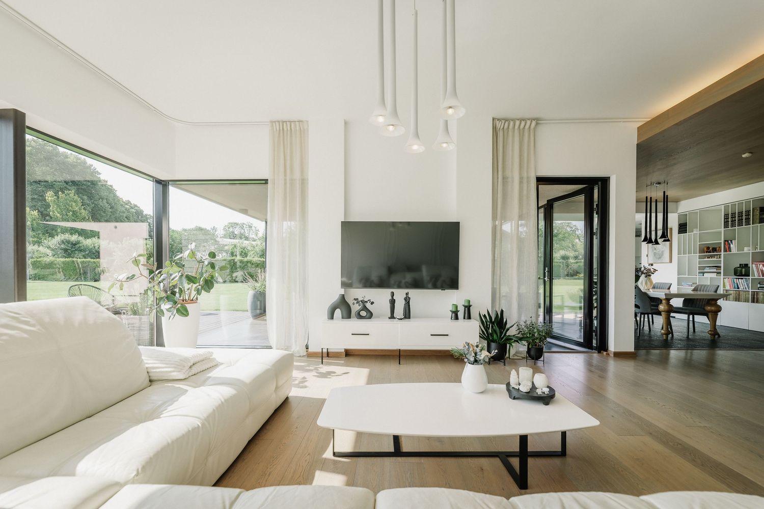 Modern white living room with leather sofa, wood floors, TV console, pendant lights, and large windows overlooking green garden.