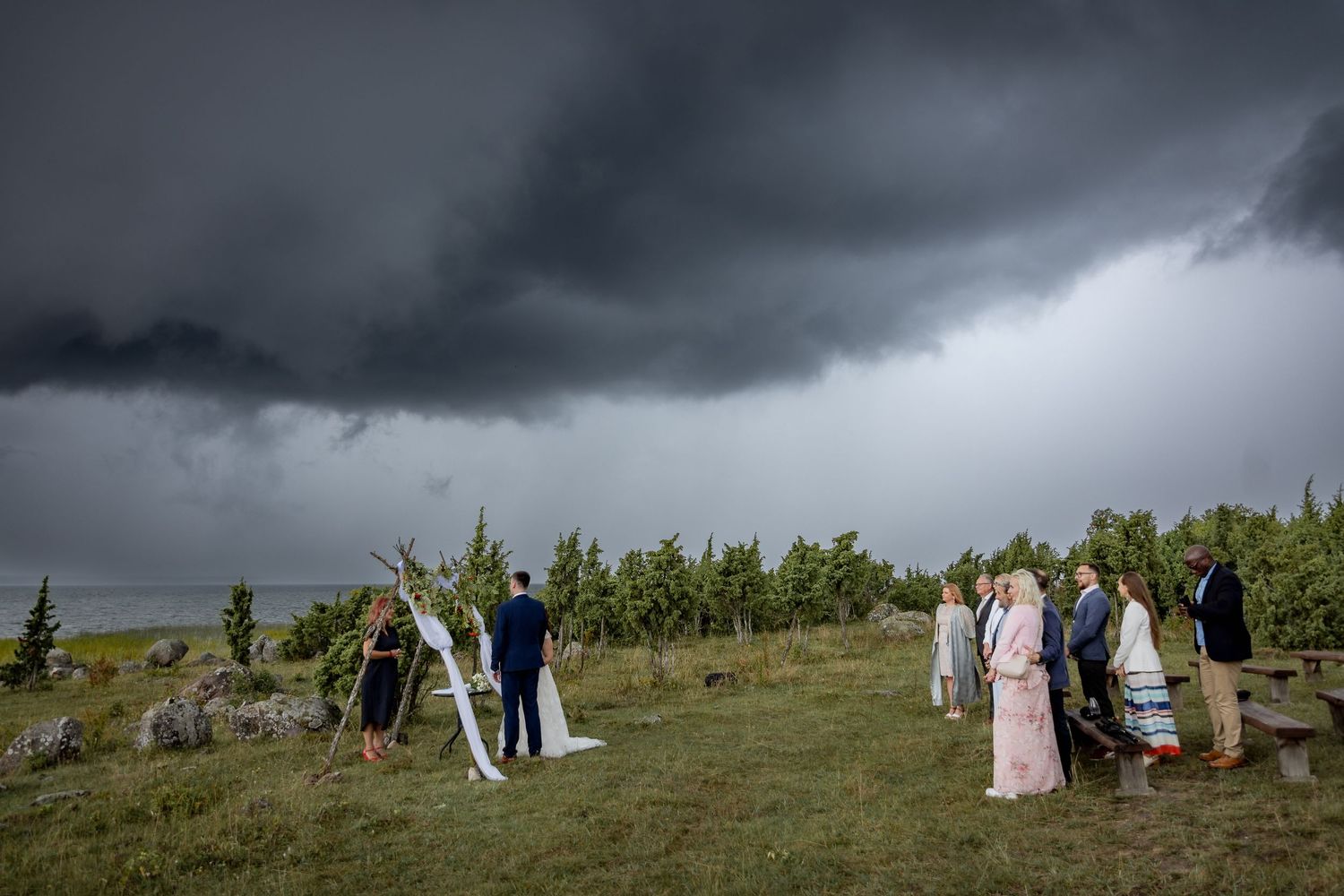 Outdoor wedding ceremony under dramatic storm clouds with guests gathered on a green hillside near a vineyard and water.